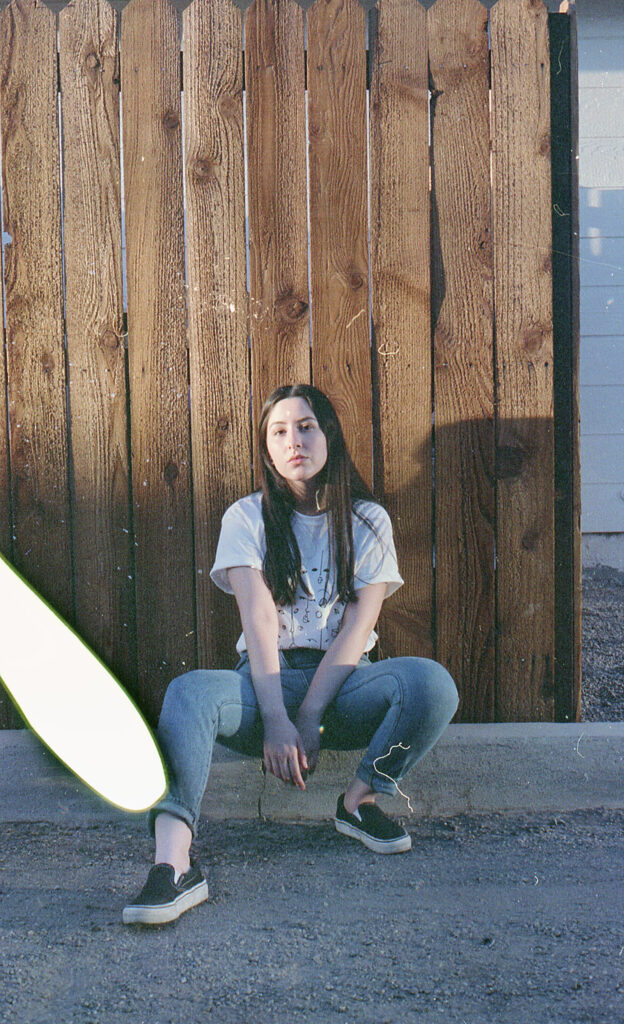 Film Photo of Gabriela Inzunza sitting against a wooden fence wearing a white t-shirt and blue jeans with a green light streak across the frame.